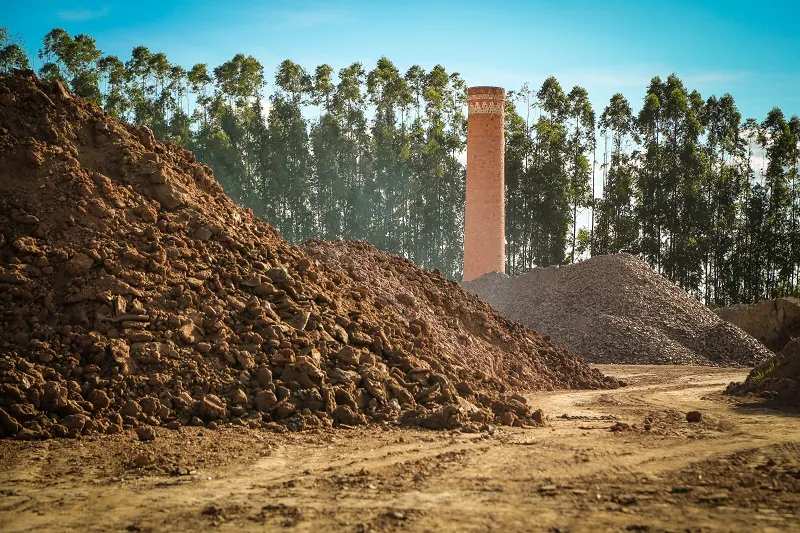 Montes de argila no pátio da fábrica, com uma chaminé de tijolos e eucaliptos ao fundo
